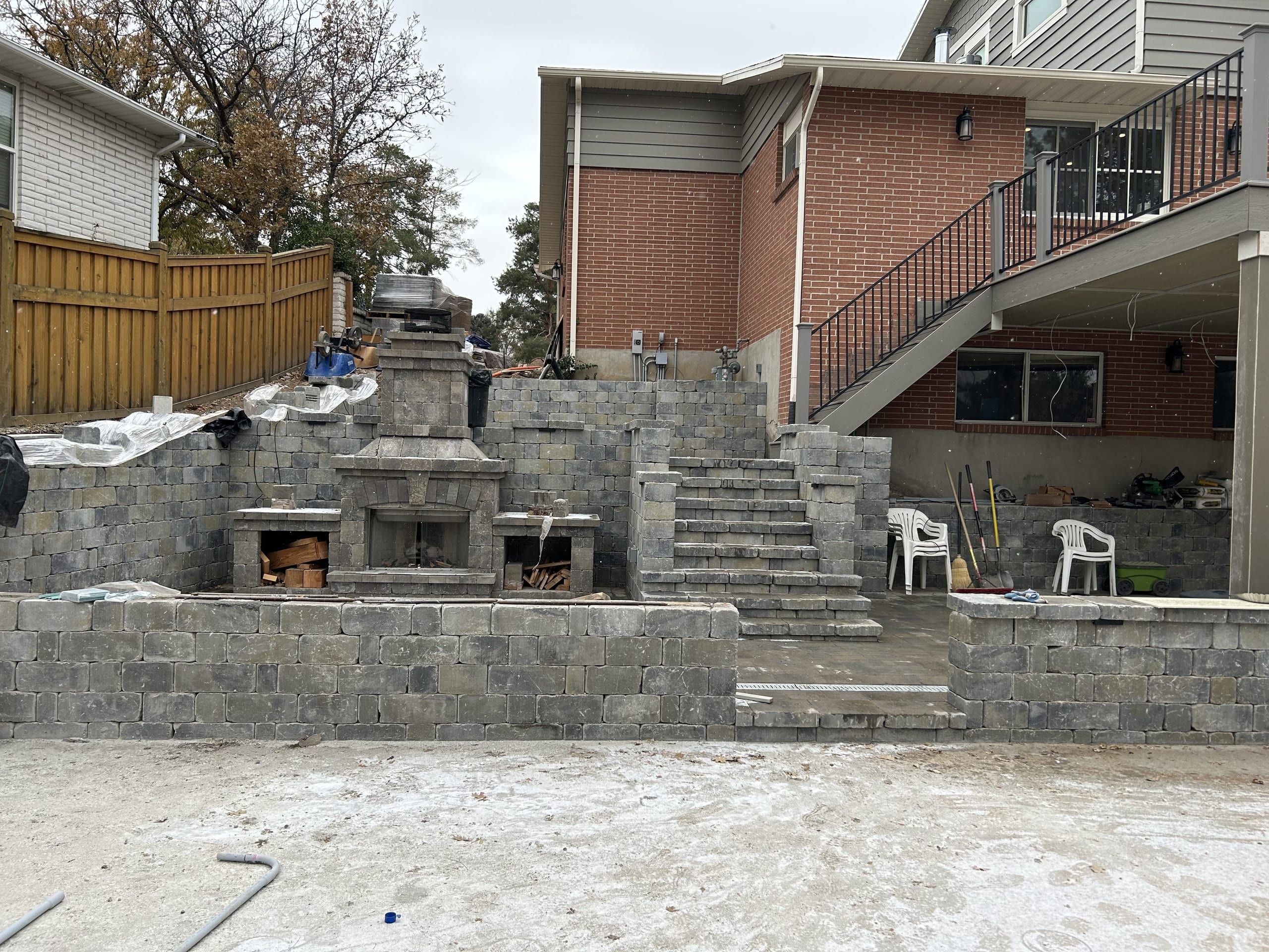 Block retaining walls, stairs and an outdoor fireplace with firewood nooks being built behind a brick house