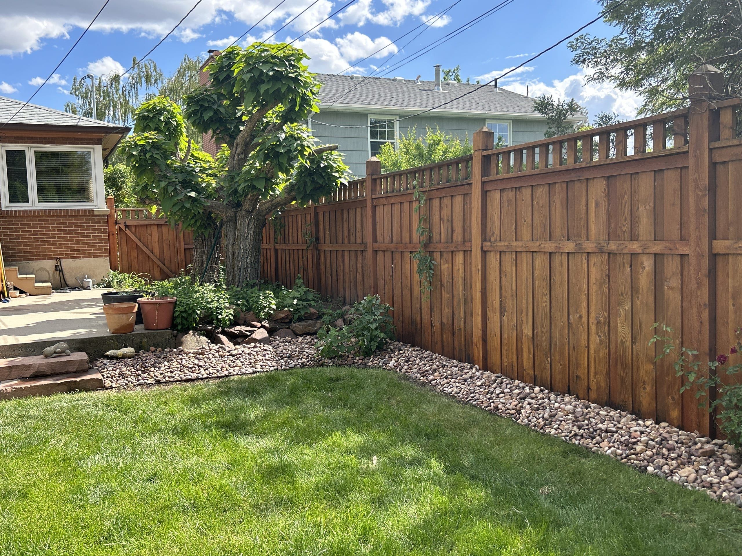 Tall reddish-brown wood privacy fence with spaced pickets along the top, edged by a river-rock border beside a green lawn, with a large tree in the corner bed