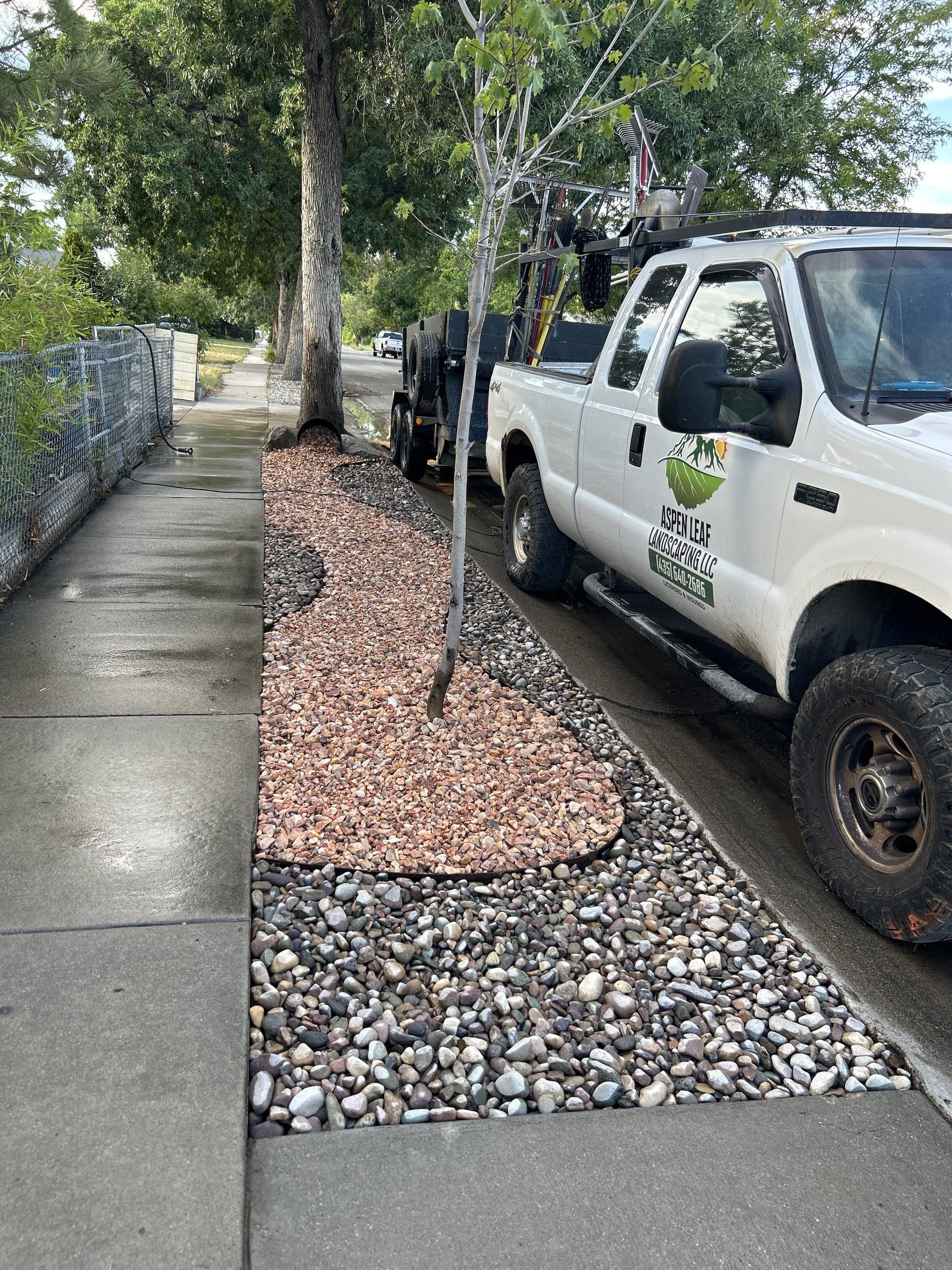 Aspen Leaf Landscaping pickup parked at the curb beside a park strip of reddish crushed rock bordered by river rock around a young tree