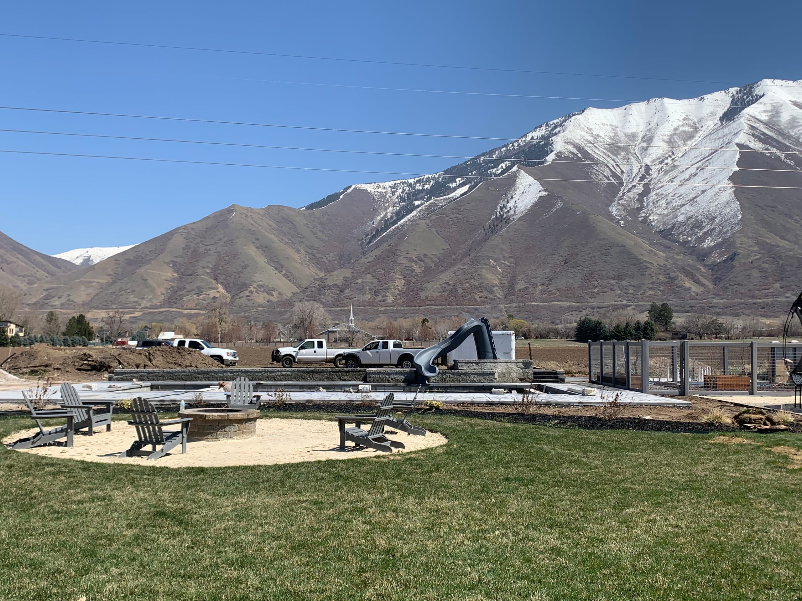 Gray Adirondack chairs around a round block fire pit on a light-colored circular pad in a lawn, with snow-capped mountains behind
