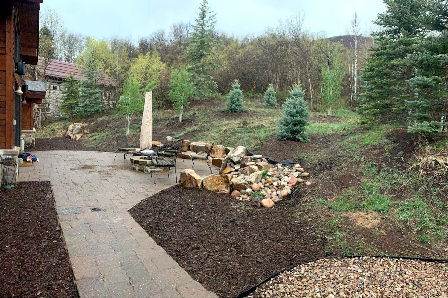Paver patio with a round fire pit and metal chairs beside boulders and river rock, with mulch beds in front and small evergreens on the slope behind
